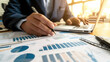 © Fareedoh - Close-up of a businessman's hands analyzing and pointing at financial charts and graphs on paper, with a laptop in the background.