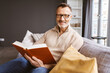 © contrastwerkstatt - Middle-aged Man with Glasses Sitting Relaxingly on Sofa, Holding a Book, and Smiling at the Camera