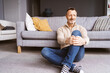© contrastwerkstatt - Senior Man Sitting Relaxed on the Floor in Front of the Sofa, Holding His Knee and Looking to the Side