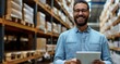 A hardware warehouse is the setting for a laughing, smiling salesman who stands while reviewing supplies on his tablet.