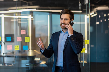 Mature businessman talking joyfully on the phone in the middle of the office, man in a business suit smiling standing by the window at the workplace, boss financier professional.