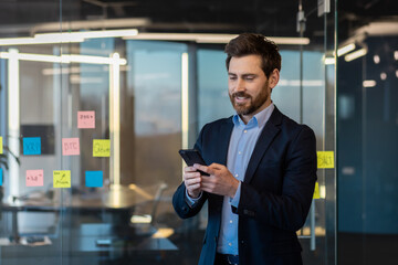 A businessman in a business suit is satisfied with the results of the achievement with a phone in his hands, a male boss is using an application on a smartphone, reading a message and dialing a call.