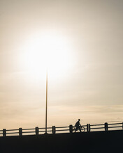 Man Walking With His Bicycle Beside Him On Top Of The Bridge, With The Sun In The Background. Sunbeams Shining Beneath The Silhouette Of The Man During His Solitary Walk And The Bridge.




