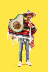 Wall Mural - Happy young Mexican man in sombrero, poncho and with avocado pinata showing thumb-up on yellow background