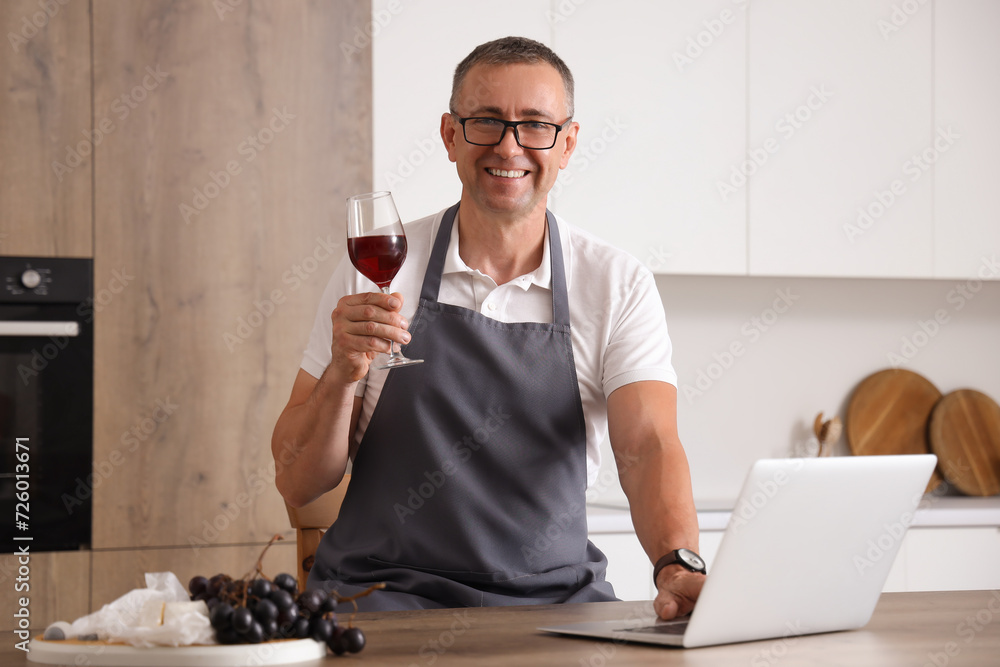 Mature man with glass of red wine using laptop in kitchen