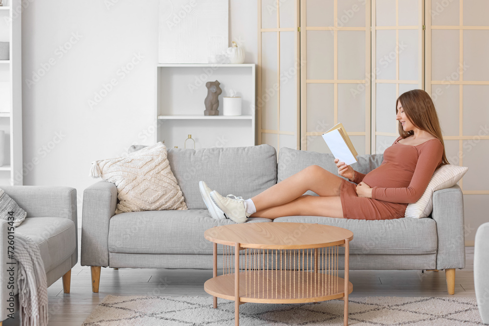 Young pregnant woman reading book on sofa at home