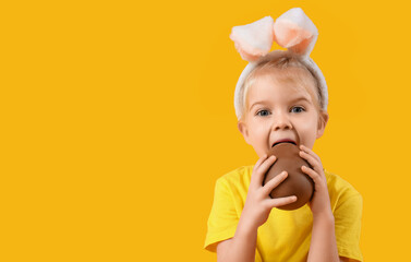 Wall Mural - Cute little girl in bunny ears eating chocolate egg on yellow background. Easter celebration