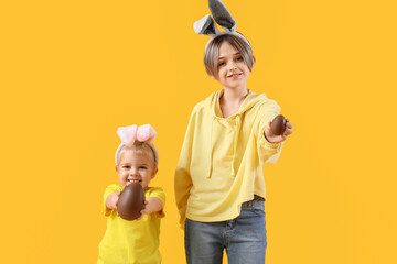 Poster - Cute little children in bunny ears with chocolate eggs on yellow background. Easter celebration