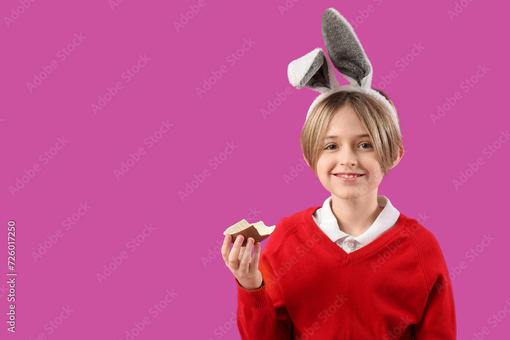 Cute little boy in bunny ears eating chocolate egg on purple background. Easter celebration