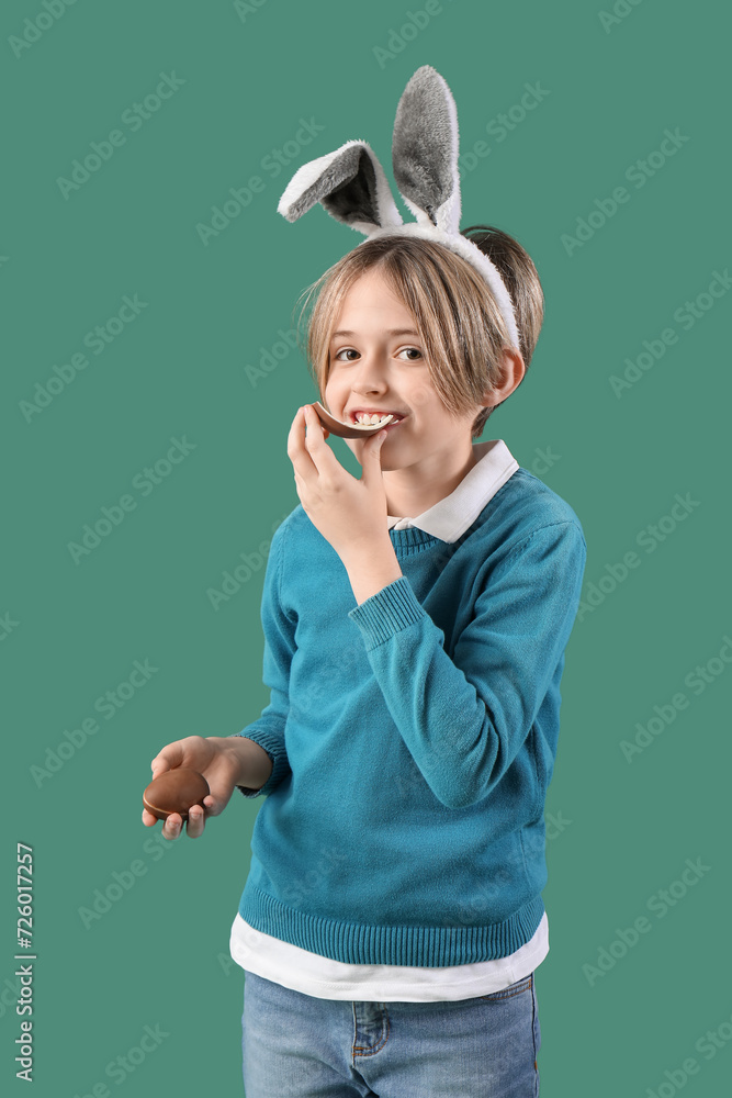 Cute little boy in bunny ears eating chocolate egg on green background. Easter celebration