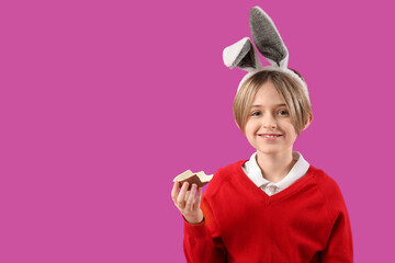 Sticker - Cute little boy in bunny ears eating chocolate egg on purple background. Easter celebration