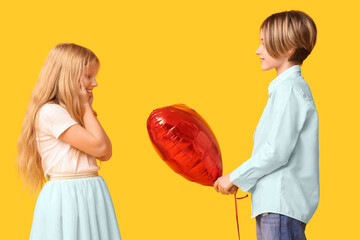 Poster - Little boy giving heart-shaped balloon to girl on yellow background. Valentine's Day celebration