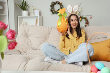 Poster - Young woman in bunny ears with Easter gift egg sitting on sofa at home