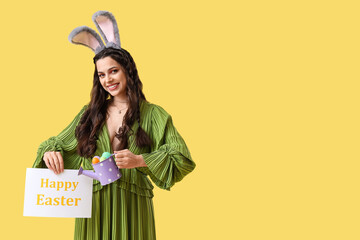 Canvas Print - Beautiful woman in bunny ears holding card with text HAPPY EASTER and eggs on yellow background