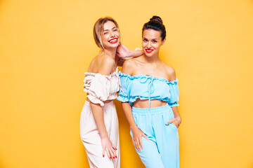 Two young beautiful smiling brunette hipster female in trendy summer clothes. Sexy carefree women posing near yellow wall in studio. Positive models having fun. Cheerful and happy