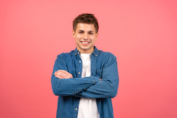 Young caucasian smiling man with arms crossed pink background