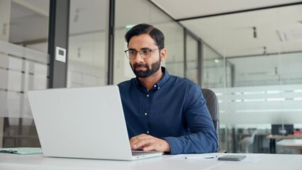 Busy Indian professional business man employee using laptop working in office. Professional financial analyst typing on computer technology working with ai solutions sitting at office desk.