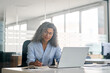 © insta_photos - Busy mature middle aged business woman, older senior female professional executive writing notes elearning online working on laptop computer device sitting at desk in office. View through glass.