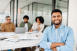 © Vadim Pastuh - Cheerful Indian professional in a denim shirt stands with arms crossed, with a bright smile, while his colleagues work together in the background, exemplifying a positive and energetic office culture