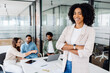 © Vadim Pastuh - Smart high-skilled young Brazilian woman with a cheerful smile stands with arms crossed on the forefront in a modern office setting with her colleagues collaborating in the background, team synergy