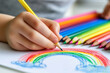 © Nataliia_Trushchenko - Little child hands draw colorful rainbow and sun shapes on white paper with a pencil. Color pencils on light blue table background. Pastel color. Closeup. Point of view shot. Children creativity time.