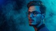 © Luiri Art - Profile of young handsome man with facial hair, in blue frame glasses looks to the side against a smokey blue background.