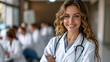 © Ash - Confident smiling female medical professional in white coat with stethoscope, standing in seminar room with colleagues in the background.