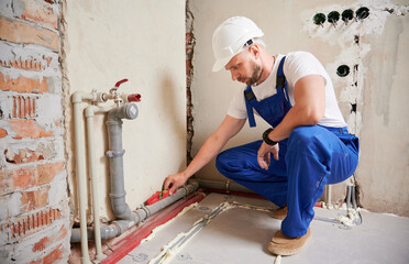 Wall Mural - Male plumber in safety construction helmet using spirit level tool while installing pipe system at home. Man measuring pipe level with building instrument in apartment under renovation.