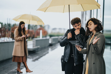 Canvas Print - Two people under yellow umbrellas in a cityscape engrossed in a serious business discussion with a tablet, ignoring the rain.