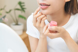 Fototapeta  - Lip care concept, happy asian young woman finger touching lips in front of mirror at home after applying lipstick balm on dry mouth from natural beauty product, skincare routine, makeup and cosmetics.