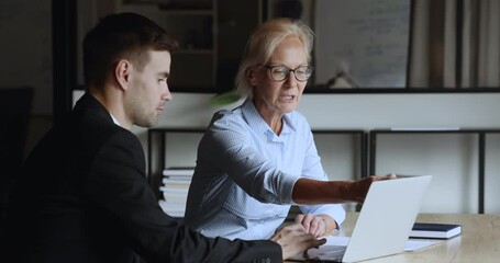 Canvas Print - Serious elder and younger colleagues discussing online project at laptop, talking at computer, looking and pointing at screen, sitting at workplace together, cooperating on startup