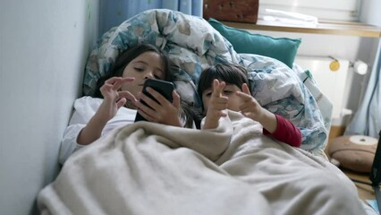 Wall Mural - Children sharing cellphone screen in bed under bedsheets. Little brother and sister using modern technology device, engaged with entertainment media online, two kids watching content
