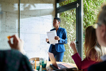 Black Woman Wearing Office Suit Holding Paper And Giving Business Presentation For Colleagues In Sunlit Room, Glass Wall On Background. Female Executive Reporting On Company Strategy