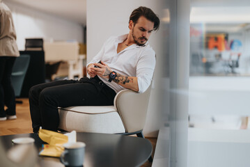 Wall Mural - A pensive young man with tattoos sits in a stylish office lounge, looking at his smart watch, embodying modern business casual style.