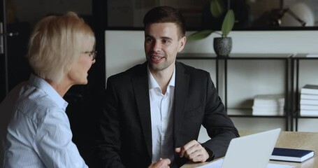 Canvas Print - Confident young manager man selling online project, startup to mature business owner woman. Younger and elder colleagues talking at workplace with laptop, shaking hands, smiling