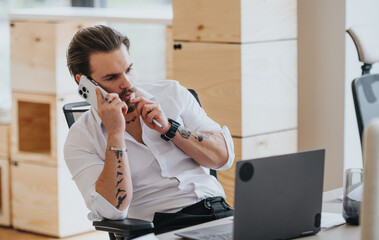 Wall Mural - A tattooed male person in a stylish office multitasks with a phone call and laptop work, exuding focus and determination.