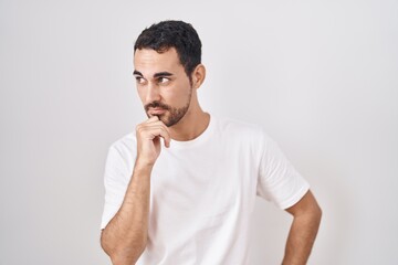 Poster - Handsome hispanic man standing over white background with hand on chin thinking about question, pensive expression. smiling with thoughtful face. doubt concept.