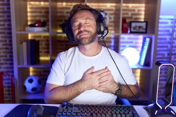 Poster - Middle age man with beard playing video games wearing headphones smiling with hands on chest with closed eyes and grateful gesture on face. health concept.