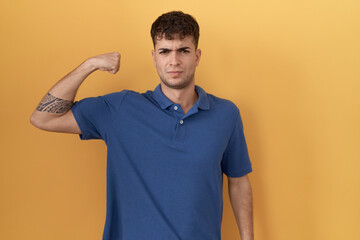 Wall Mural - Young hispanic man standing over yellow background strong person showing arm muscle, confident and proud of power