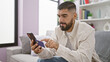 © Krakenimages.com - A bearded man in a casual shirt attentively uses a smartphone in a modern living room setting, conveying a sense of technology in everyday life.