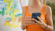 © Krakenimages.com - A young woman in an orange top uses a smartphone in a creative studio space with artwork in the background.
