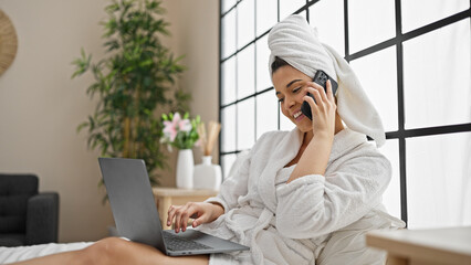 Poster - Young beautiful hispanic woman wearing bathrobe using laptop talking on smartphone at bedroom