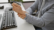 © Krakenimages.com - A hispanic man in an office wears a grey shirt, holding his wrist, denoting possible pain or injury, next to a computer keyboard.