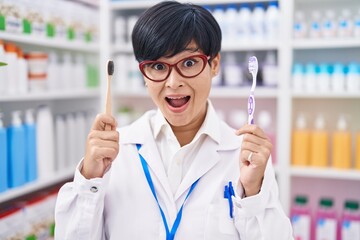 Sticker - Young asian woman with short hair doing toothbrush comparative at pharmacy celebrating crazy and amazed for success with open eyes screaming excited.