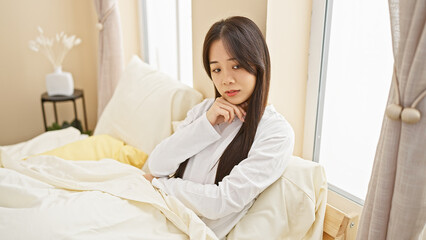 Canvas Print - A young asian woman in a white shirt sitting thoughtfully on a bed in a well-lit bedroom interior