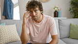 Fototapeta  - A thoughtful young hispanic man sits on a grey couch in a cozy living room, giving off a contemplative vibe.