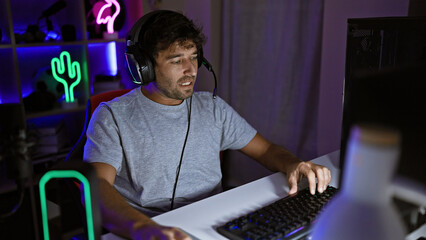 Canvas Print - Focused man gaming in a dark room with neon lights, headphones on, intensely looking at the computer screen.