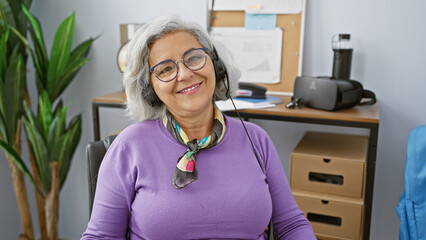 Poster - A smiling grey-haired woman wearing a headset sits in a modern office space, exuding professionalism and warmth.