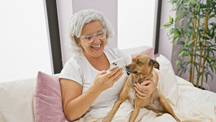 Poster - A joyful mature woman using a smartphone cuddles with her dog on a bed inside a cozy room.