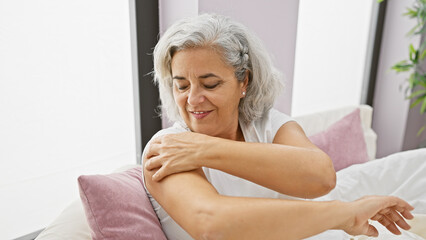 Poster - Mature woman applying lotion on her arm in a cozy bedroom setting, showcasing self-care and domestic tranquility.